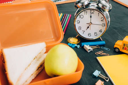 selective focus of lunch box with tasty sandwiches and ripe apple near vintage alarm clock and school supplies on black chalkboardの写真素材
