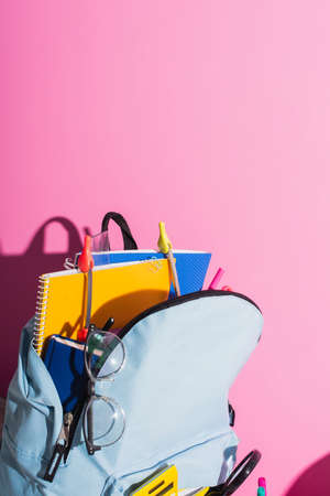 blue backpack with notebooks, school supplies and eyeglasses on pink