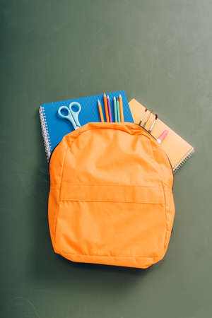 top view of yellow backpack with copy books, scissors, color pencils and paintbrushes on green...