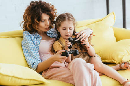 KYIV, UKRAINE - JUNE 19, 2020: young babysitter and adorable kid sitting on yellow sofa with joysticksの写真素材