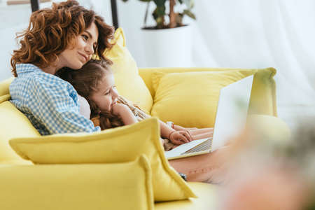 selective focus of smiling babysitter and child sitting on sofa and using laptopの写真素材
