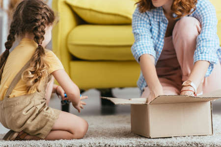 cropped view of babysitter and kid sitting on floor near cardboard box
