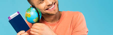 panoramic crop of happy bearded man holding passport and globe isolated on blueの写真素材