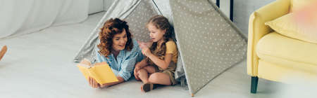 high angle view of smiling nanny reading book to adorable kid in play tent, panoramic shotの写真素材