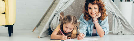 happy nanny looking at camera while drawing together with child in play tent, panoramic shotの写真素材