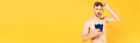 panoramic crop of shocked and shirtless man touching sunglasses and holding passports isolated on yellowの写真素材