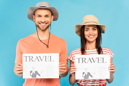 happy couple in hats holding travel newspapers isolated on blueの写真素材