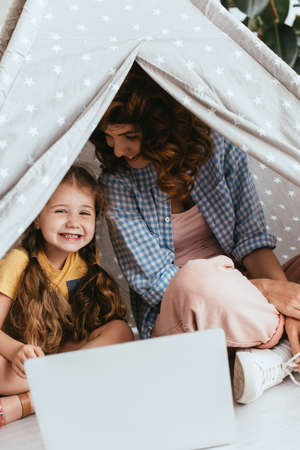 happy babysitter and adorable child sitting in toy wigwam near laptop