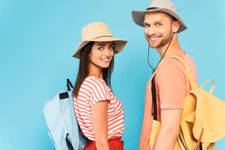 happy couple in hats with backpacks looking at camera isolated on blueの写真素材