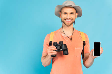 cheerful man holding binoculars and smartphone with blank screen on blue