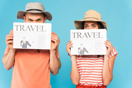 couple in hats covering faces with travel newspapers isolated on blueの写真素材