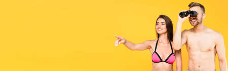 website header of cheerful woman pointing with finger while shirtless man looking through binoculars isolated on yellowの写真素材