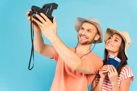 happy man in hat holding vintage camera and taking selfie with cheerful girl isolated on blue