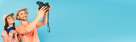 panoramic shot of happy man in hat holding vintage camera and taking selfie with cheerful girl isolated on blueの写真素材