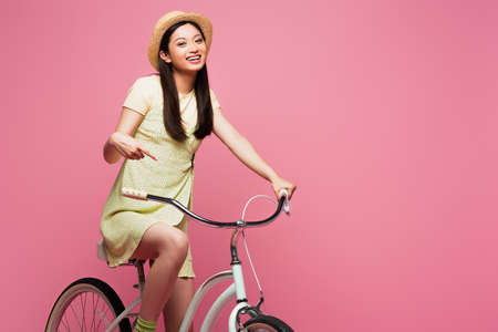 smiling asian young woman in straw hat riding bicycle isolated on pink