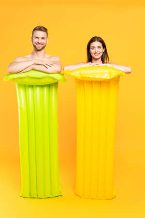 happy couple looking at camera and standing near inflatable mattresses on yellow