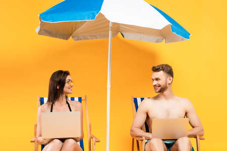 happy couple with laptops sitting on deck chairs near beach umbrella on yellowの写真素材