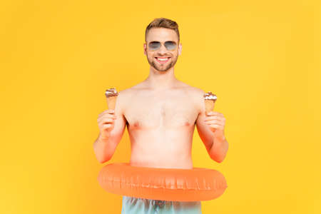 cheerful and shirtless man in sunglasses with inflatable ring holding ice cream cones isolated on yellowの写真素材