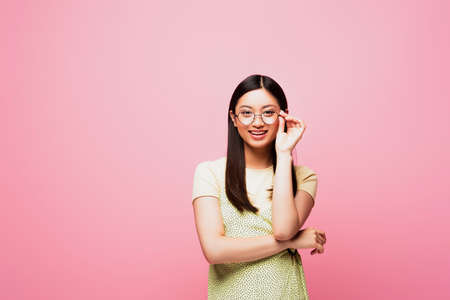 happy asian girl touching glasses while looking at camera isolated on pink