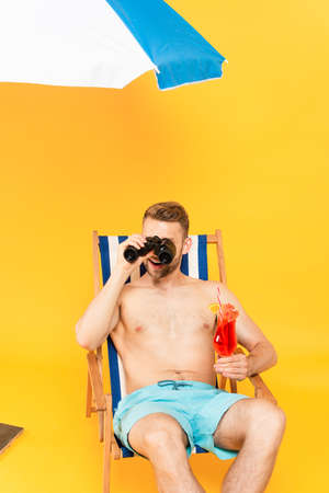 happy shirtless man holding cocktail and looking through binoculars on yellowの写真素材