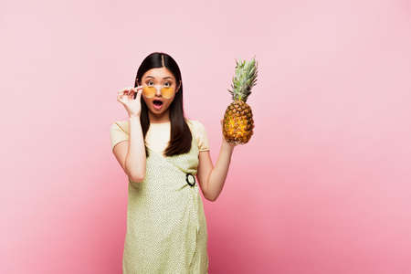 shocked asian girl in sunglasses holding ripe pineapple on pinkの写真素材