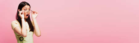 panoramic shot of shocked asian girl looking away and touching glasses isolated on pinkの写真素材