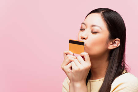 young asian woman with closed eyes kissing credit card isolated on pink