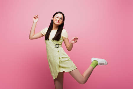 cheerful and young asian woman in glasses jumping on pink
