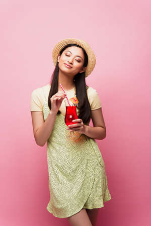 smiling young asian woman with closed eyes standing and holding glass with cocktail on pinkの写真素材