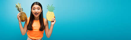 panoramic concept of excited asian girl holding ripe and carton pineapples on blueの写真素材