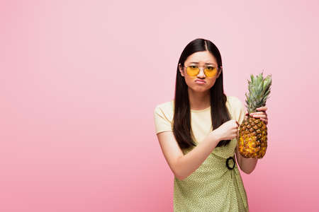 sad asian woman in sunglasses holding fresh pineapple isolated on pink