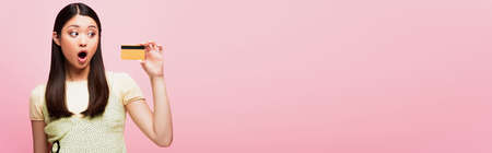 panoramic crop of shocked young asian woman holding credit card isolated on pink