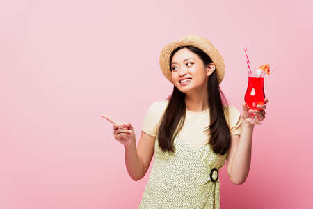smiling young asian woman in straw hat holding glass with cocktail and pointing with finger isolated on pinkの写真素材