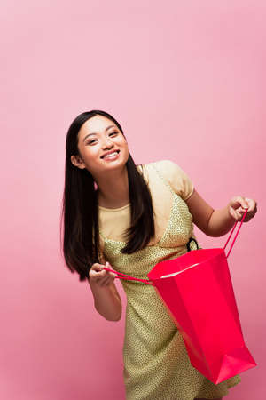 smiling young asian woman holding shopping bag isolated on pink