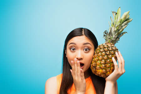 shocked asian girl covering mouth and holding ripe pineapple on blueの写真素材
