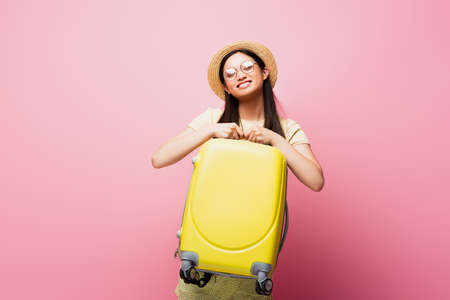 happy asian girl in glasses and straw hat holding yellow luggage on pinkの写真素材