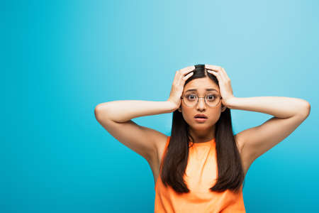 shocked asian woman in glasses touching head on blue