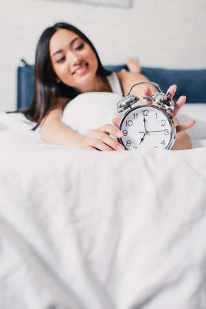 Selective focus of smiling asian woman holding alarm clock on bed at morning