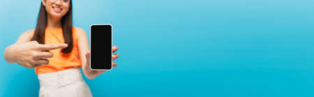 panoramic crop of happy girl pointing with finger at smartphone with blank screen isolated on blue