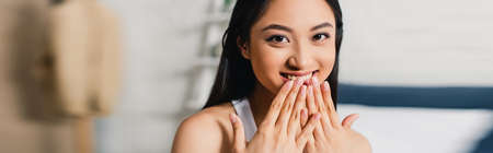 Panoramic shot of positive asian woman with hands near mouth looking at camera at home