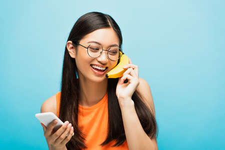 happy asian girl in glasses using smartphone and holding banana near ear on blue