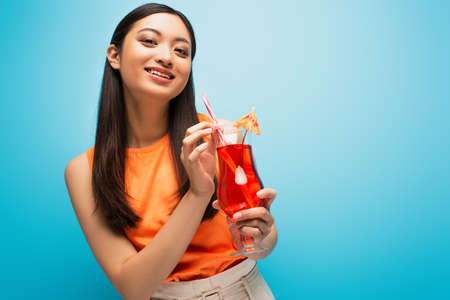 happy asian girl holding glass with cocktail on blue