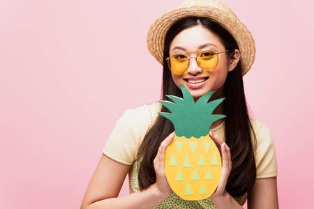 cheerful asian woman in sunglasses and straw hat holding paper pineapple isolated on pinkの写真素材