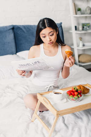Selective focus of asian woman reading newspaper and eating breakfast on bed at home