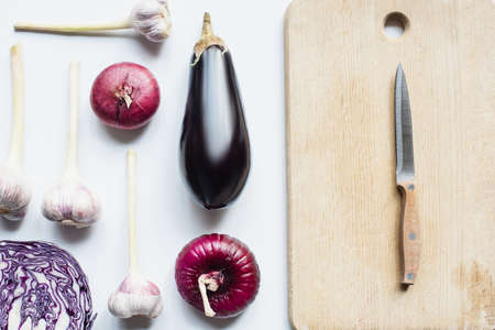 flat lay with red onion, red cabbage, eggplant and garlic near wooden cutting board with knife on white backgroundの写真素材