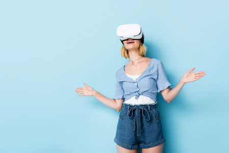 young surprised woman in virtual reality headset gesturing on blue