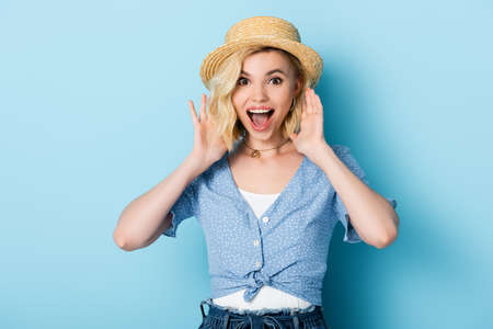 excited young woman in straw hat gesturing and looking at camera on blue