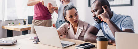 Panoramic shot of businesswoman looking at pensive african american colleague near gadgets on table...