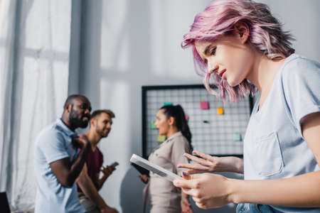 Selective focus of young businesswoman using digital tablet with multiethnic colleagues in office