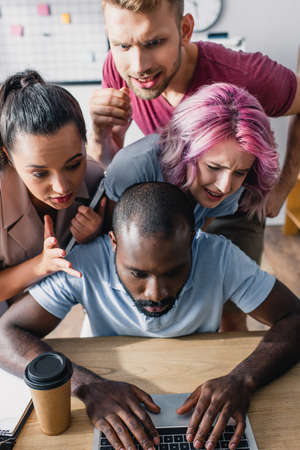 Concentrated multicultural business people looking at african american colleague using laptop at table in officeの写真素材